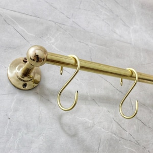 May include: A brass kitchen rail with S-shaped hooks, mounted on a marble-effect wall. The rail has a rounded end and a circular mounting plate. The rail is designed for hanging utensils.