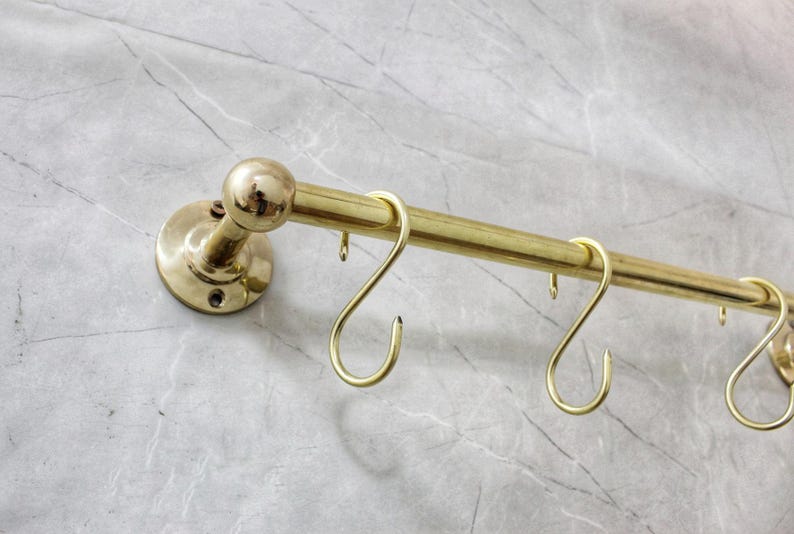 May include: A brass-colored kitchen rail with several S-shaped hooks. The rail is mounted on a marble-patterned wall. The rail has a rounded end and a circular mounting plate.