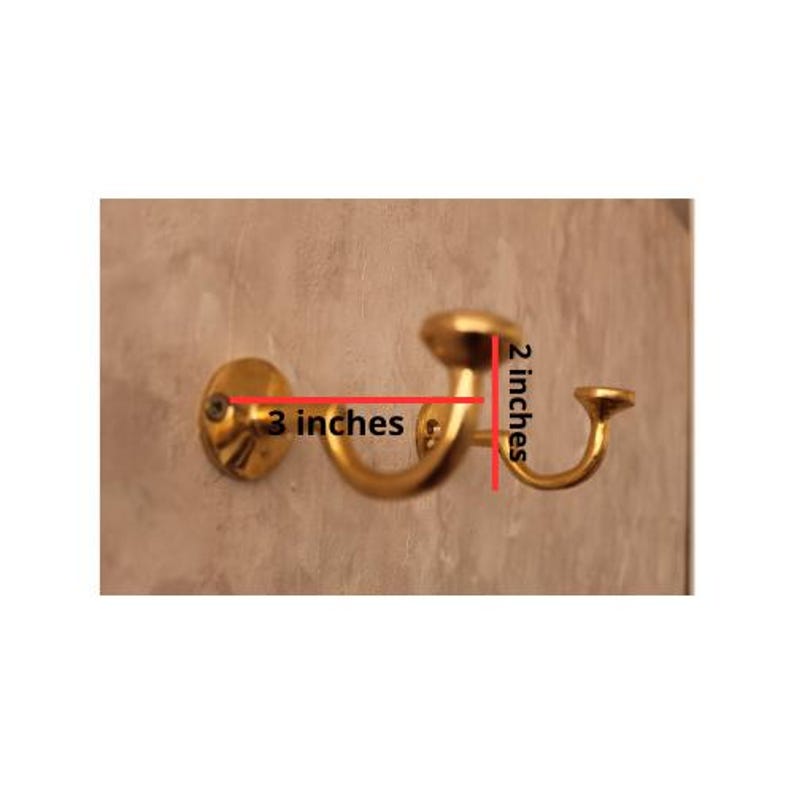May include: A gold-colored wall hook, 3 inches wide and 2 inches high, mounted on a wall. The hook has a curved design with a circular base and a rounded top. The hook is designed for hanging items.