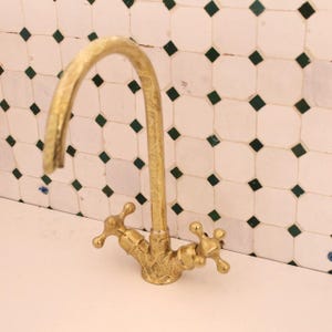 Puede incluir: Un grifo dorado con un caño curvo y manijas en forma de cruz. El grifo tiene un diseño ornamentado y se encuentra sobre un fondo de azulejos blancos con detalles de diamantes verdes.