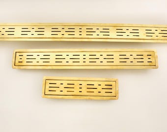 May include: Three rectangular brass shower drain covers of varying sizes. Each drain cover has a series of horizontal and vertical slots for water drainage. The brass has a warm, golden hue, and the drains are arranged against a neutral background.