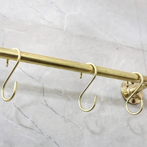 May include: A brass-colored hanging rod with several S-shaped hooks. The rod is mounted on a wall with a marble-like pattern. The hooks are evenly spaced along the rod, designed for hanging items.