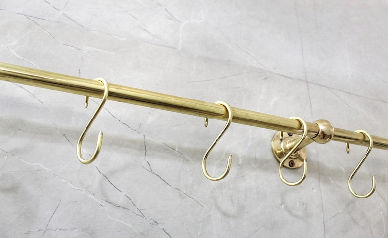 May include: A brass-colored hanging rod with several S-shaped hooks. The rod is mounted on a wall with a marble-like pattern. The hooks are evenly spaced along the rod, designed for hanging items.