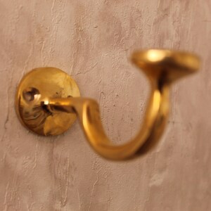 May include: A brass wall hook with a curved design. The hook is mounted on a circular base and has a polished, golden finish. The hook is designed to hold items and is mounted on a textured, light brown wall.