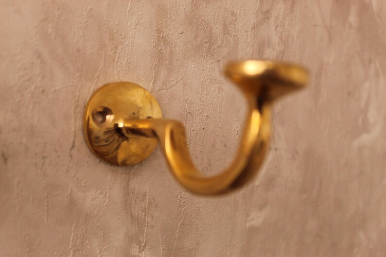 May include: A brass wall hook with a curved design. The hook is mounted on a circular base and has a polished, golden finish. The hook is designed to hold items and is mounted on a textured, light brown wall.