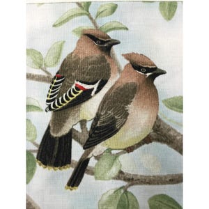 May include: A fabric print featuring two cedar waxwing birds perched on a brown tree branch with green leaves against a light blue background. The birds have brown bodies, black eye masks, and wings with white, red, and yellow markings.