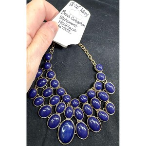 May include: A gold-tone statement necklace featuring multiple rows of smooth, oval-shaped navy blue cabochons. The attached white tag displays the handwritten text: 18-20" Navy Bead Cabachon Statement Necklace Gold Tone.
