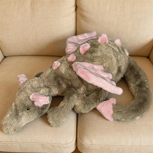 以下が含まれることがあります： グレーとピンクの色合いのぬいぐるみドラゴン。ドラゴンは長い体、尾、翼を持っています。ピンクのアクセントは、翼、耳、背中のトゲにあります。おもちゃはベージュのソファの上にあります。