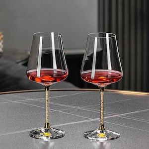 May include: Two clear wine glasses filled with red liquid. The glasses have a gold stem with a crystal-like base. The glasses are on a grey surface. The background is dark.