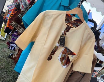 Puede incluir: Una camisa amarilla clara de manga corta con un patrón texturizado, un bolsillo estampado y ribetes. Una camisa azul es visible en el fondo, junto con otras telas estampadas coloridas. Las camisas están colgadas en un perchero.