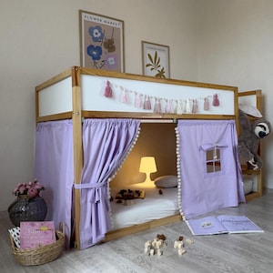 Puede incluir: Una cama alta de madera con una cortina de color lila claro y una ventana. La cama tiene un cabecero blanco y está decorada con borlas rosas y blancas. Una pequeña lámpara ilumina el espacio dentro de la cama. Un peluche de perezoso cuelga de un lado.