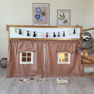 Op de afbeelding: Een houten bedframe met een roze gordijn speelhuis. Het gordijn heeft twee vensteruitsparingen met een crèmekleurige rand. Een slinger van kwasten en kralen hangt boven het gordijn. Een knuffelsluis is aan het bedframe bevestigd.