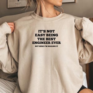 Könnte beinhalten: Beiges Sweatshirt mit Rundhalsausschnitt und dem Text "IT'S NOT EASY BEING THE BEST ENGINEER EVER BUT HERE I'M NAILING IT" in schwarzen Buchstaben. Das Sweatshirt hat eine neutrale Farbe und scheint aus weichem Material zu sein.
