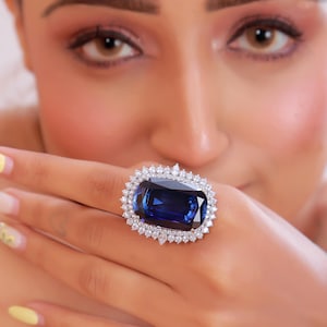 Könnte beinhalten: Ein Ring mit einem großen, rechteckigen, tiefblauen Edelstein im Kissenschliff, gefasst in ein silberfarbenes Metallband. Der zentrale blaue Stein ist von einem doppelten Kranz aus kleinen, klaren, runden weißen Steinen umgeben.