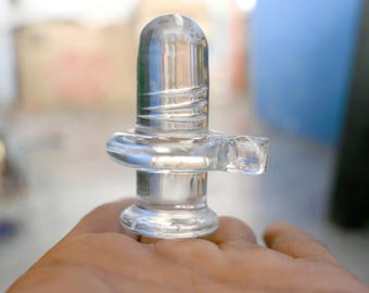 May include: A clear glass lingam statue, a religious symbol, is held in a hand. The statue has a cylindrical shape with horizontal ridges and a circular base. The background is blurred.