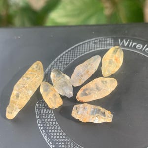 Op de afbeelding: Een verzameling ruwe, ongeslepen edelstenen in tinten geel en helder. De stenen hebben een natuurlijke, langwerpige vorm en variërende afmetingen. De edelstenen worden getoond op een donker, patroonrijk oppervlak, dat hun doorschijnende kwaliteit benadrukt.
