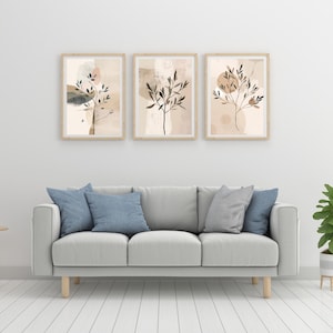 May include: Three framed abstract art prints with botanical designs in neutral tones. The artwork features beige, brown, and grey colours, with a light wood frame. The prints are displayed above a grey sofa with blue and grey cushions.