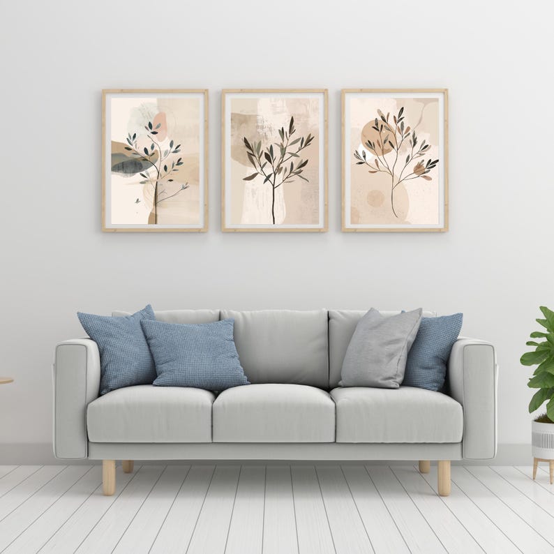 May include: Three framed abstract art prints with botanical designs in neutral tones. The artwork features beige, brown, and grey colours, with a light wood frame. The prints are displayed above a grey sofa with blue and grey cushions.