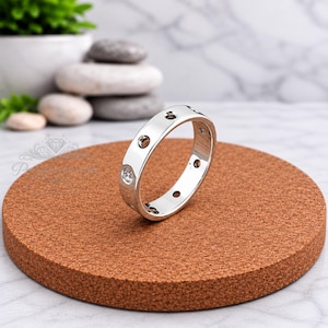 May include: A polished silver-tone metal ring featuring a series of evenly spaced circular indentations along the outer band. The ring is positioned upright on a circular brown cork surface, showcasing a smooth, reflective finish throughout.