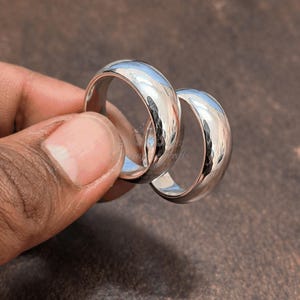 Puede incluir: Dos anillos de plata pulida con un diseño de banda redondeada. Los anillos se sostienen en una mano y reflejan la luz, creando una apariencia brillante y brillante. Los anillos son probablemente alianzas o joyería de moda.
