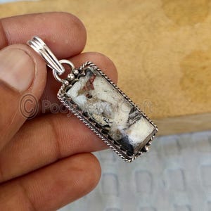 May include: A rectangular pendant with a silver-colored bail. The pendant features a stone with a mix of white, black, and reddish-brown patterns, set within a decorative silver frame. The frame has a twisted rope design.