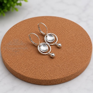 May include: A pair of silver-toned earrings featuring a lever-back closure, a circular ring frame, a central hammered concave disc, and a small dangling spherical bead at the bottom. The earrings have a polished metallic finish and a geometric design.