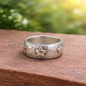 Puede incluir: Un anillo plateado con una banda con estrellas en relieve. El anillo se coloca sobre una superficie de madera con un fondo verde borroso, creando un contraste entre el brillo metálico y los tonos naturales. El diseño del anillo es simple pero llamativo.