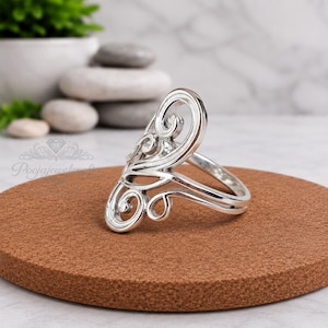 Könnte beinhalten: Ein silberner Ring mit einem länglichen, durchbrochenen Design und geschwungenen, schnörkelartigen Mustern. Das Metall hat eine polierte, reflektierende Oberfläche und die Struktur besteht aus ineinandergreifenden Schleifen und Kurven.