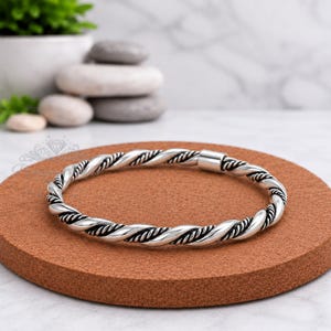 Puede incluir: Una pulsera de tono plateado con un diseño de cuerda trenzada, acabado pulido y ranuras oxidadas oscuras. El brazalete circular tiene un cierre metálico cilíndrico y liso que une los dos extremos de la banda de metal trenzada.