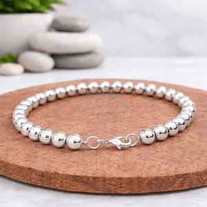 May include: A silver beaded bracelet with a lobster clasp. The bracelet is made of round, shiny beads. The bracelet is displayed on a brown surface, with a blurred background of white stones and greenery.