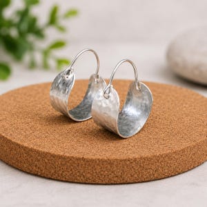 May include: A pair of silver-toned hoop earrings featuring a wide, curved band with a textured, hammered finish. Each earring is constructed with a thin, rounded wire loop that connects to the main body, creating a distinct open-hoop silhouette.