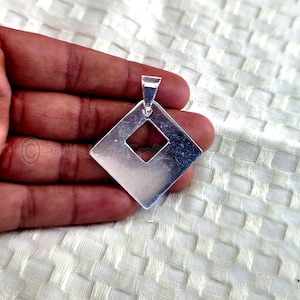 May include: A silver pendant featuring a diamond shape with a central diamond cut-out. The pendant has a loop for a chain. The jewellery is held in a hand against a white background.