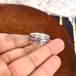 Puede incluir: Un anillo giratorio plateado con una banda de estrellas y lunas crecientes. El anillo se sostiene en una mano, mostrando el diseño celestial. El diseño del anillo está grabado en la banda, creando una pieza de joyería detallada y llamativa.