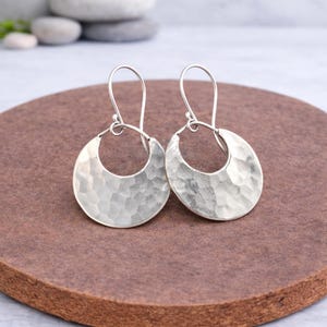 Può includere: Un paio di orecchini a cerchio argentati con texture martellata. Gli orecchini hanno una forma a mezzaluna con finitura martellata e sono sospesi a un semplice gancio. Gli orecchini sono esposti su una superficie marrone.
