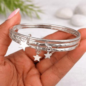 May include: A set of three silver bangles with a hammered texture. Each bangle has a small star charm dangling from it. The bangles are held in a hand, with a blurred background.
