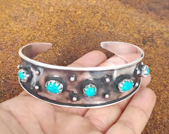 May include: Silver cuff bracelet with turquoise stones. The bracelet has a wide band with a textured surface and is adorned with five oval turquoise stones and small silver accents. The open-ended design allows for easy wear.