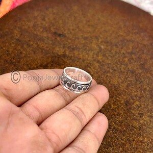 May include: A silver ring with a wave pattern design. The ring is held in a hand and has a band with a repeating black wave pattern. The ring is a simple band style.