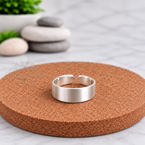May include: A silver-coloured metal ring with a smooth, matte finish and a simple open-band design. The ring features a flat, uniform surface and a clean, circular shape, showcasing a minimalist aesthetic with no additional patterns or engravings.