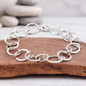 May include: A silver bracelet with a series of interconnected, hammered-look oval and circular links. The bracelet is secured with a lobster clasp. The jewellery is displayed on a reddish-brown surface.