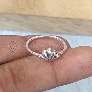 Puede incluir: Un anillo de plata con diseño de concha. El anillo presenta una banda delicada y una pieza central detallada en forma de concha. El anillo se muestra en un dedo, destacando su tamaño y diseño. Es una joya.