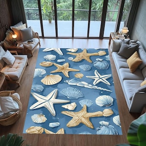 Puede incluir: Una alfombra rectangular azul con ilustraciones de conchas marinas y estrellas de mar en blanco, beige y dorado. La alfombra está en un salón con luz natural y muebles, creando un ambiente costero.