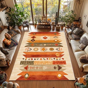 Puede incluir: Una alfombra rectangular grande con un fondo color crema y un patrón geométrico en tonos naranja, marrón y verde. El diseño incluye motivos de diamantes, flechas y rayas, creando una estética del suroeste o tribal. La alfombra está colocada sobre un suelo de madera.