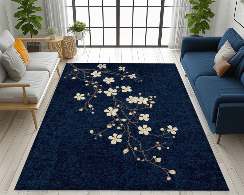 May include: A navy blue area rug featuring a floral branch design with white blossoms and buds. The rug is rectangular and placed on a light wood floor, with a sofa and window in the background.