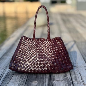 May include: A burgundy-colored tote bag with a woven design. The bag has two braided handles and a trapezoidal shape. The bag is made of a shiny material and sits on a wooden surface.