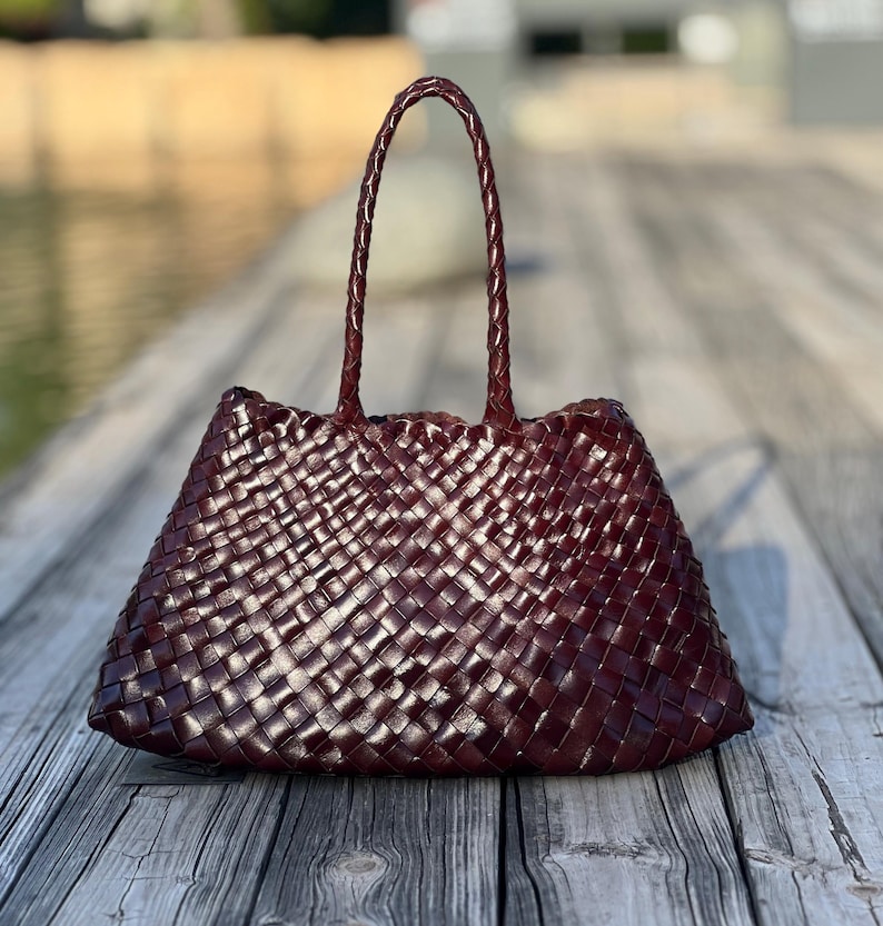 May include: A burgundy-colored tote bag with a woven design. The bag has two braided handles and a trapezoidal shape. The bag is made of a shiny material and sits on a wooden surface.