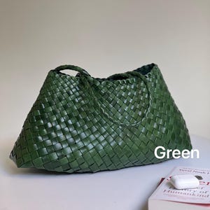 May include: A green woven tote bag with two handles. The bag is made of a tightly woven material and has a trapezoid shape. The word "Green" is written on the bag. A book and a pair of white wireless earbuds are next to the bag.