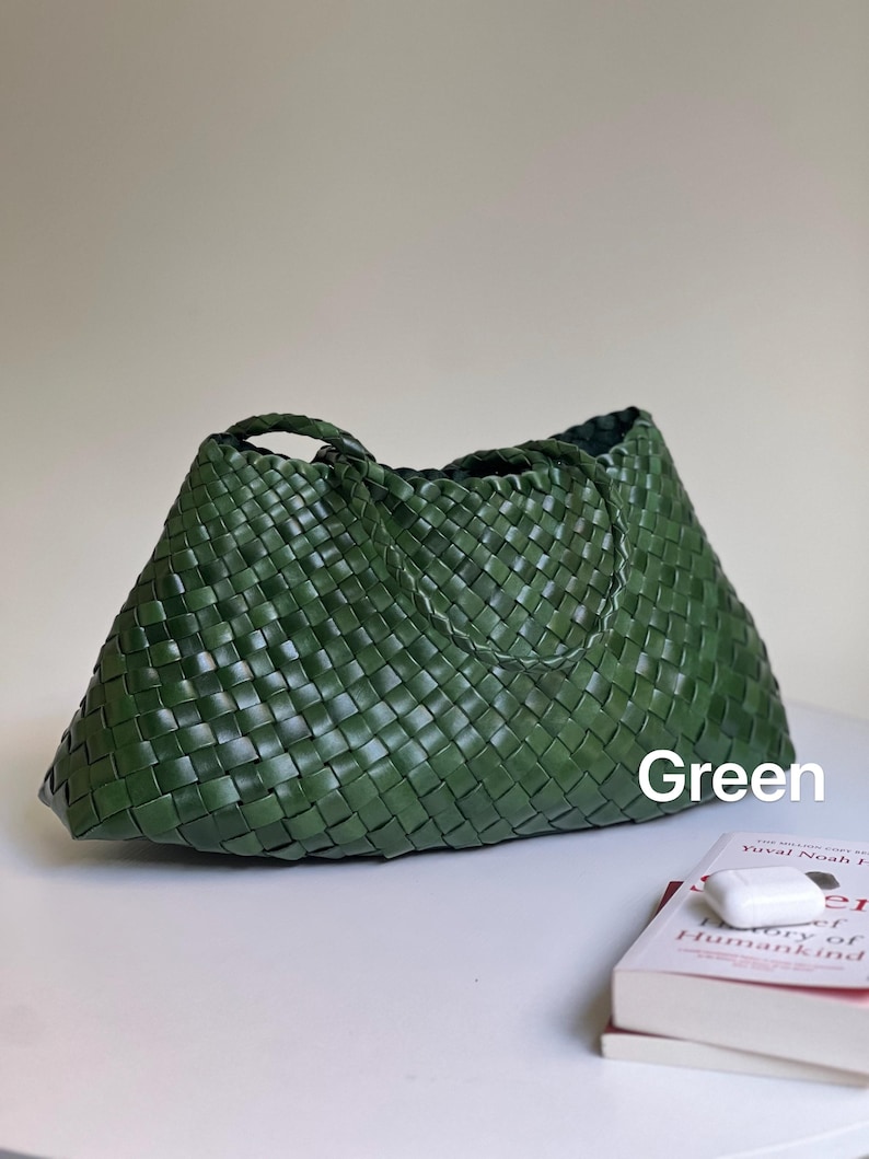 May include: A green woven tote bag with two handles. The bag is made of a tightly woven material and has a trapezoid shape. The word "Green" is written on the bag. A book and a pair of white wireless earbuds are next to the bag.