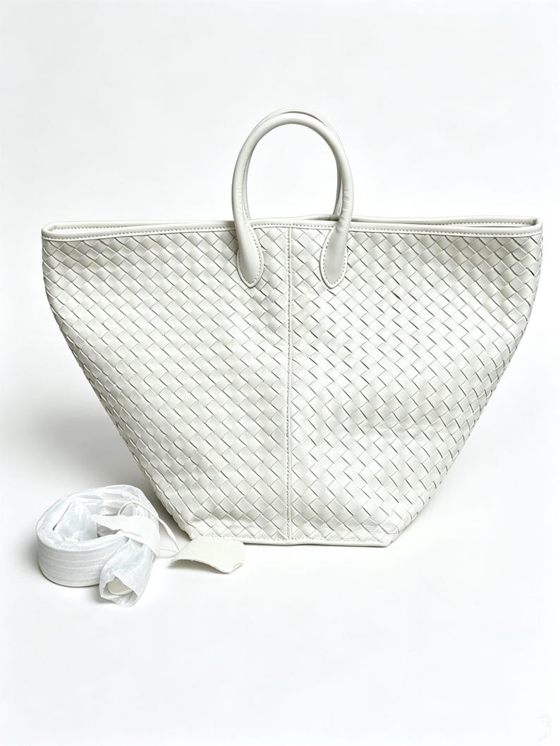 May include: A white woven leather tote bag with two top handles and a detachable shoulder strap. The bag has a trapezoid shape and a textured surface. The bag is made of white leather.
