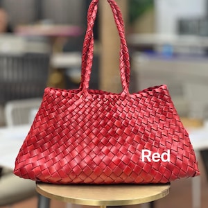 May include: A red woven tote bag with braided handles. The bag is rectangular with a trapezoidal shape and sits on a gold-colored table. The word "Red" is printed on the bag.