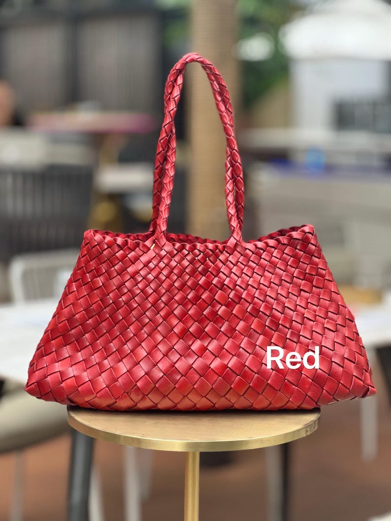 May include: A red woven tote bag with braided handles. The bag is rectangular with a trapezoidal shape and sits on a gold-colored table. The word "Red" is printed on the bag.